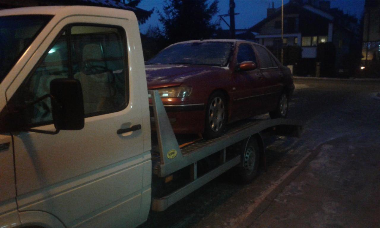 Czerwony samochód osobowy zabezpieczony na platformie lawety, widoczny fragment białego pojazdu holowniczego, zimowa sceneria z budynkami w tle.