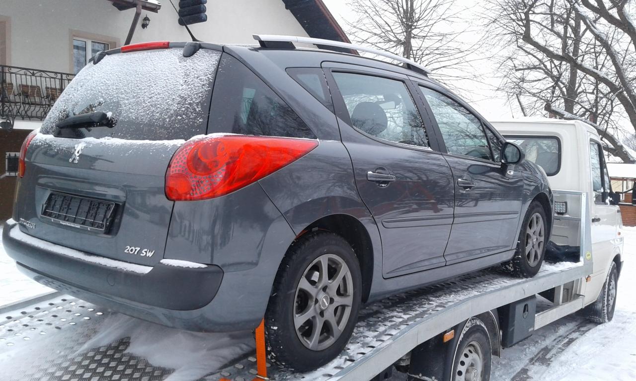 Szary samochód Peugeot 207 SW pokryty śniegiem, transportowany na lawecie w zimowy dzień.