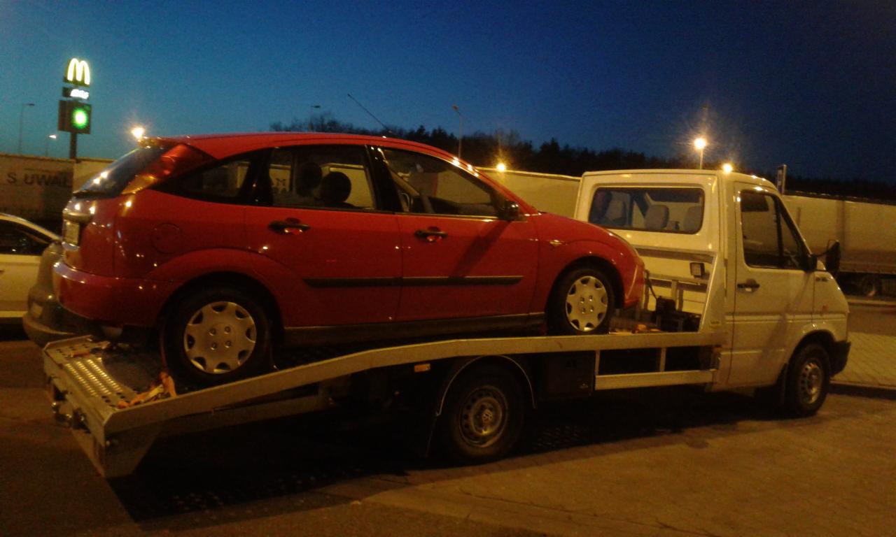 Czerwony samochód osobowy marki Ford Focus przewożony na białej lawecie przy drodze, w tle widoczny szyld McDonald's.