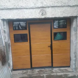 Nowa brama garażowa segmentowa z okienkami w kolorze drewna, czarne ramy, widoczny fragment starej, otwartej bramy obok, ściana garażu z kamerą.