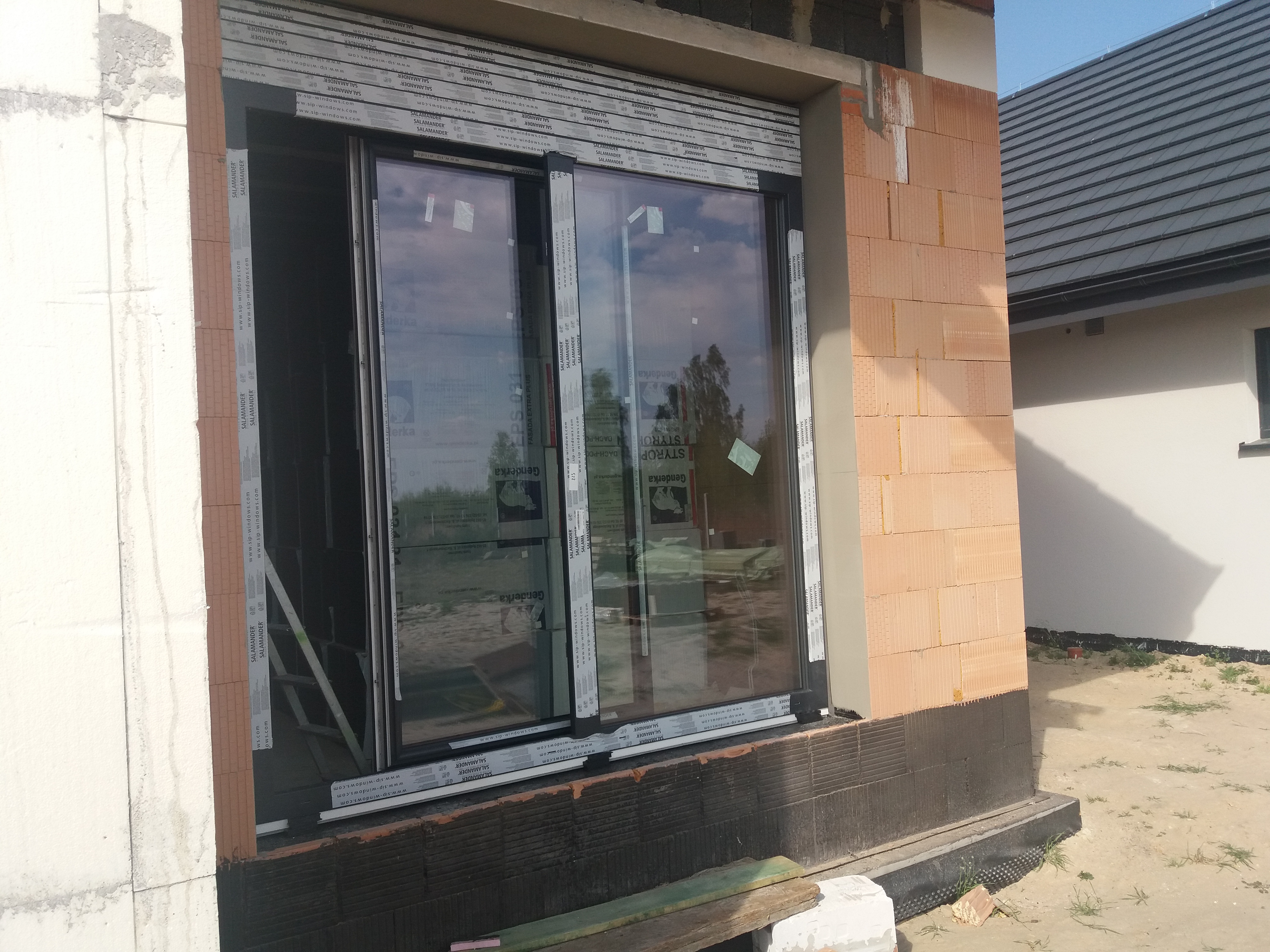 Montaż przesuwnych okien z profili Salamander w surowym stanie budynku z cegły ceramicznej, widoczne folie ochronne na ramach.