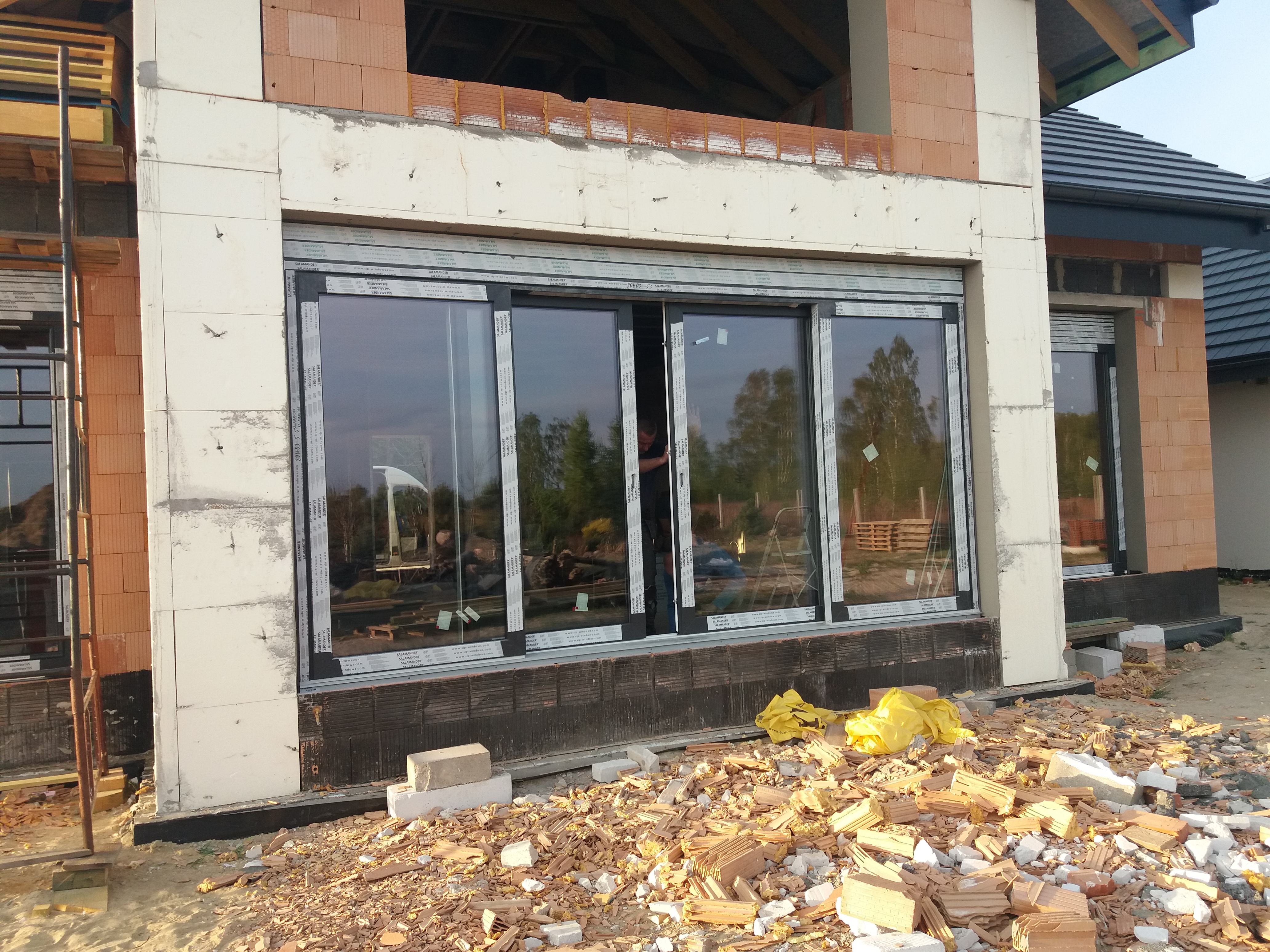 Montaż trzyskrzydłowych okien przesuwnych w surowym stanie budynku, widoczne piankowane ocieplenie i gruz budowlany na zewnątrz.