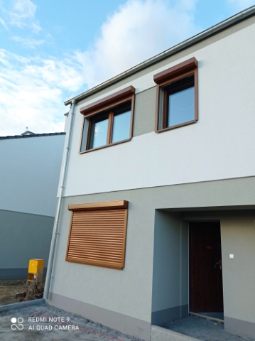 Elewacja domu z trzema oknami z brązowymi roletami zewnętrznymi, dwie z nich zwinięte, jedna zasłonięta, widok z dołu na tle błękitnego nieba.