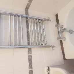 Zamontowany chromowany grzejnik łazienkowy na ścianie z białych płytek z dekoracyjnym pasem, widoczna bateria wannowa i fragment wanny oraz umywalki.