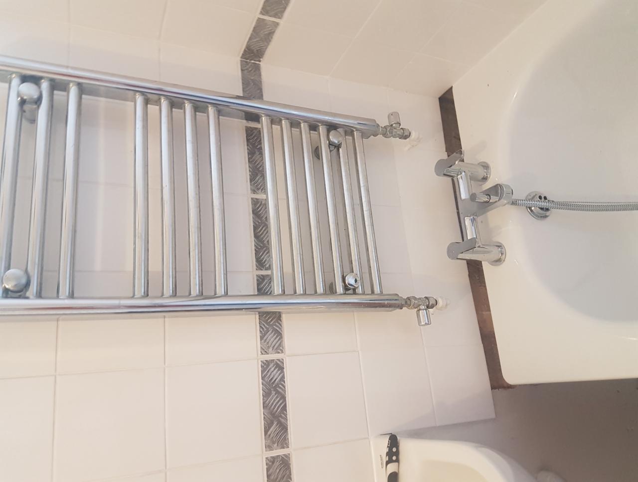 Zamontowany chromowany grzejnik łazienkowy na ścianie z białych płytek z dekoracyjnym pasem, widoczna bateria wannowa i fragment wanny oraz umywalki.