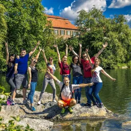 Grupa uśmiechniętych osób pozuje na skale nad jeziorem, w tle zieleń i budynek z czerwoną dachówką, błękitne niebo z chmurami. Uczestnicy imprezy integracyjnej.