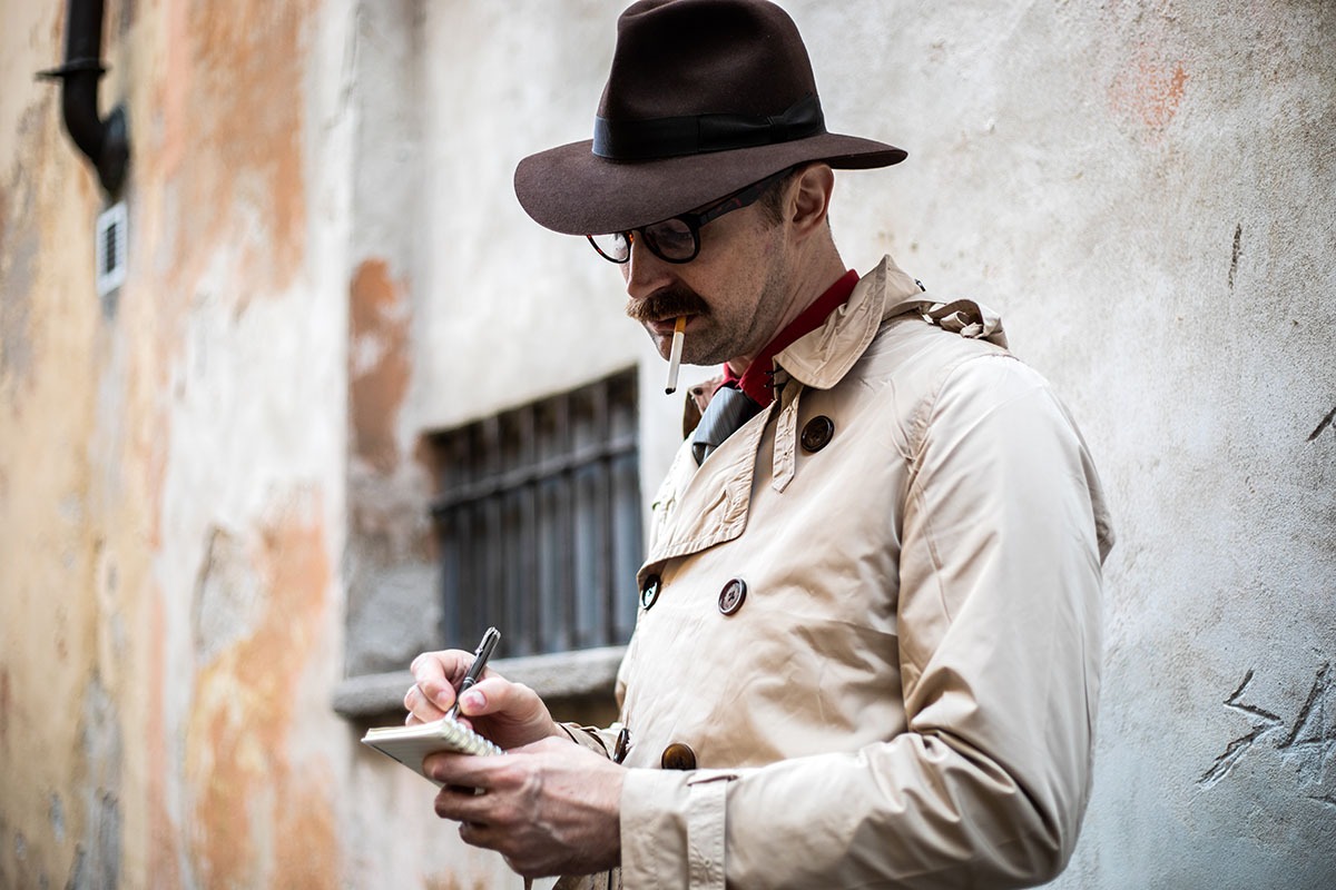 Mężczyzna w kapeluszu i płaszczu pisze w notatniku, paląc papierosa na tle teksturowanej ściany z oknem w tle. Kadr od pasa w górę.