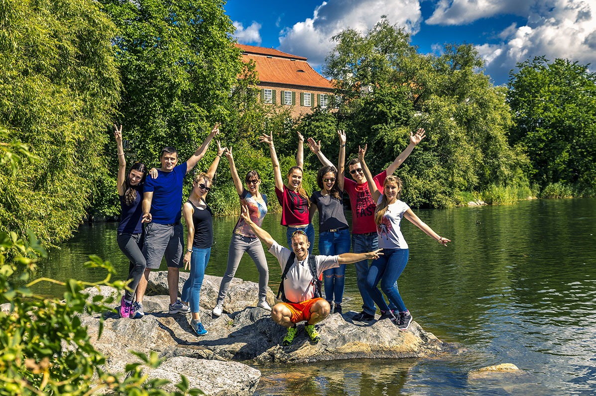 Grupa uśmiechniętych osób pozuje na skale nad jeziorem, w tle zieleń i budynek z czerwoną dachówką, błękitne niebo z chmurami. Uczestnicy imprezy integracyjnej.