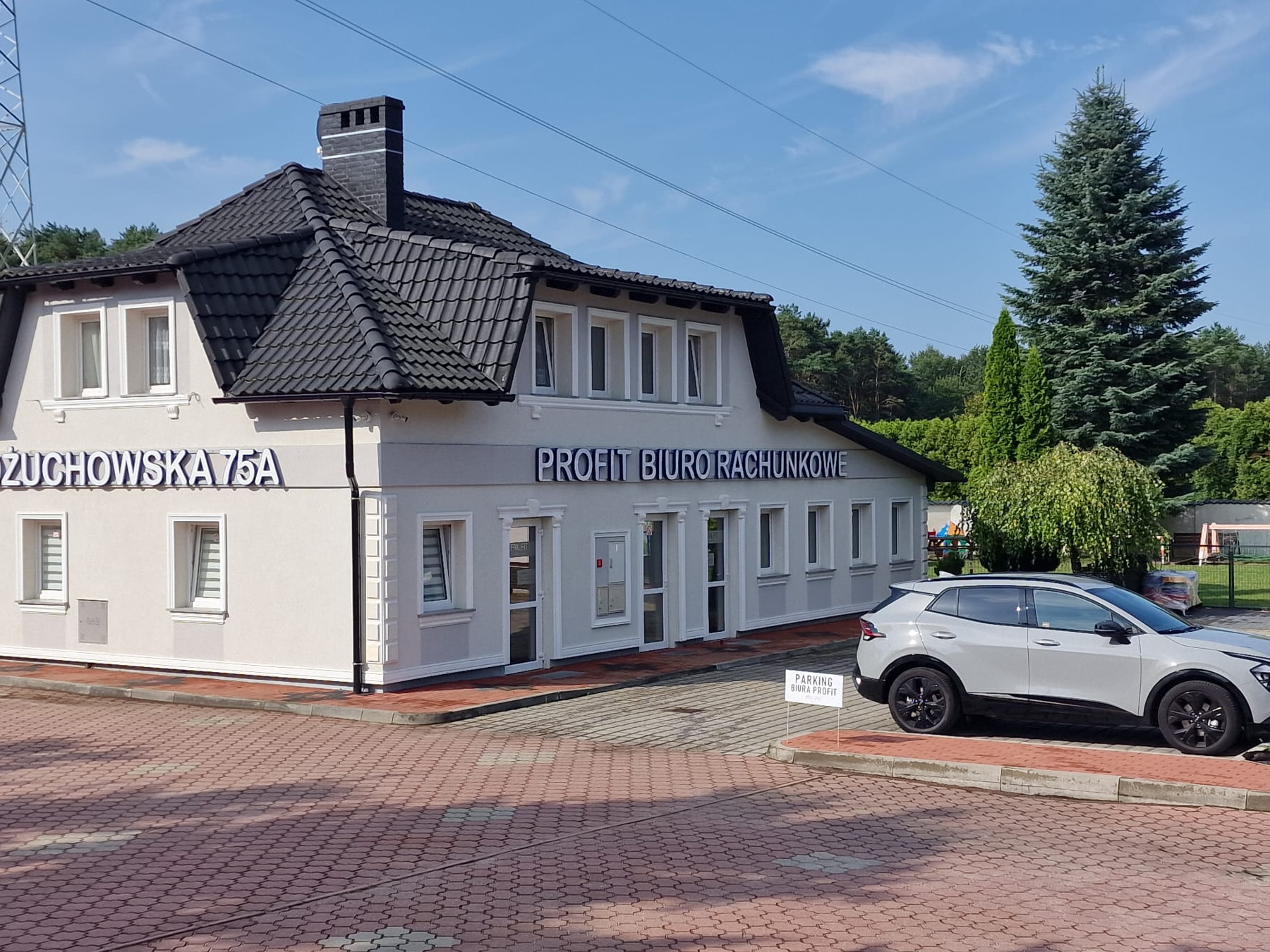 Budynek biura rachunkowego z napisem 'PROFIT BIURO RACHUNKOWE' i adresem 'ŻUCHOWSKA 75A', obok zaparkowany srebrny samochód, w tle zieleń drzew i błękitne niebo.