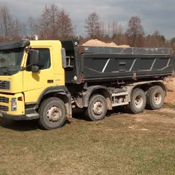 Żółta ciężarówka Volvo FM 400 z czarną skrzynią ładunkową wypełnioną piaskiem stoi na trawiastym terenie w słoneczny dzień, na drugim planie widoczne drzewa i fragment hałdy.
