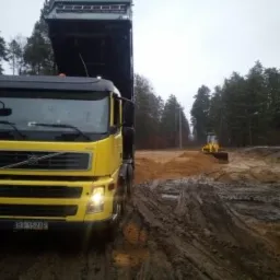 Żółta ciężarówka Volvo z podniesioną skrzynią ładunkową stoi na błotnistym terenie budowy, w tle widoczny spychacz wyrównujący teren.