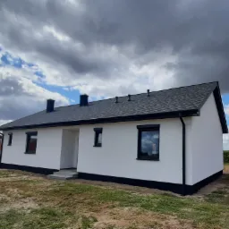 Nowoczesny dom jednorodzinny z białą elewacją i ciemną podmurówką, czarnymi ramami okiennymi i dachem z gontu bitumicznego, widoczne kominy i rynny, pochmurne niebo w tle.