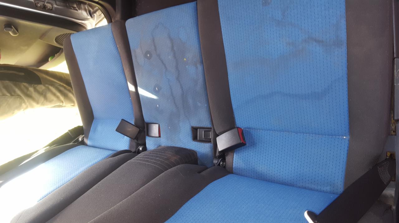 Brudna, niebieska tapicerka tylnej kanapy samochodu przed czyszczeniem, widoczne plamy i zacieki, pasy bezpieczeństwa.