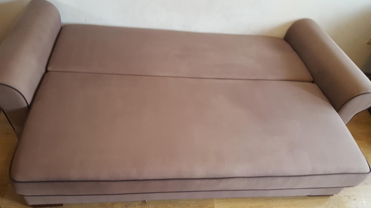 Rozłożona, beżowa kanapa z tapicerką i ciemnobrązową lamówką, widoczna z góry, na drewnianej podłodze.