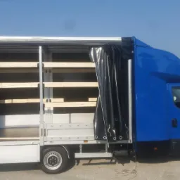 Niebieski samochód dostawczy z otwartą przestrzenią ładunkową wyposażoną w drewniane półki, częściowo zasłoniętą czarną plandeką, zaparkowany przed budynkiem z garażem.