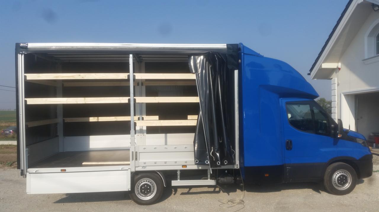 Niebieski samochód dostawczy z otwartą przestrzenią ładunkową wyposażoną w drewniane półki, częściowo zasłoniętą czarną plandeką, zaparkowany przed budynkiem z garażem.