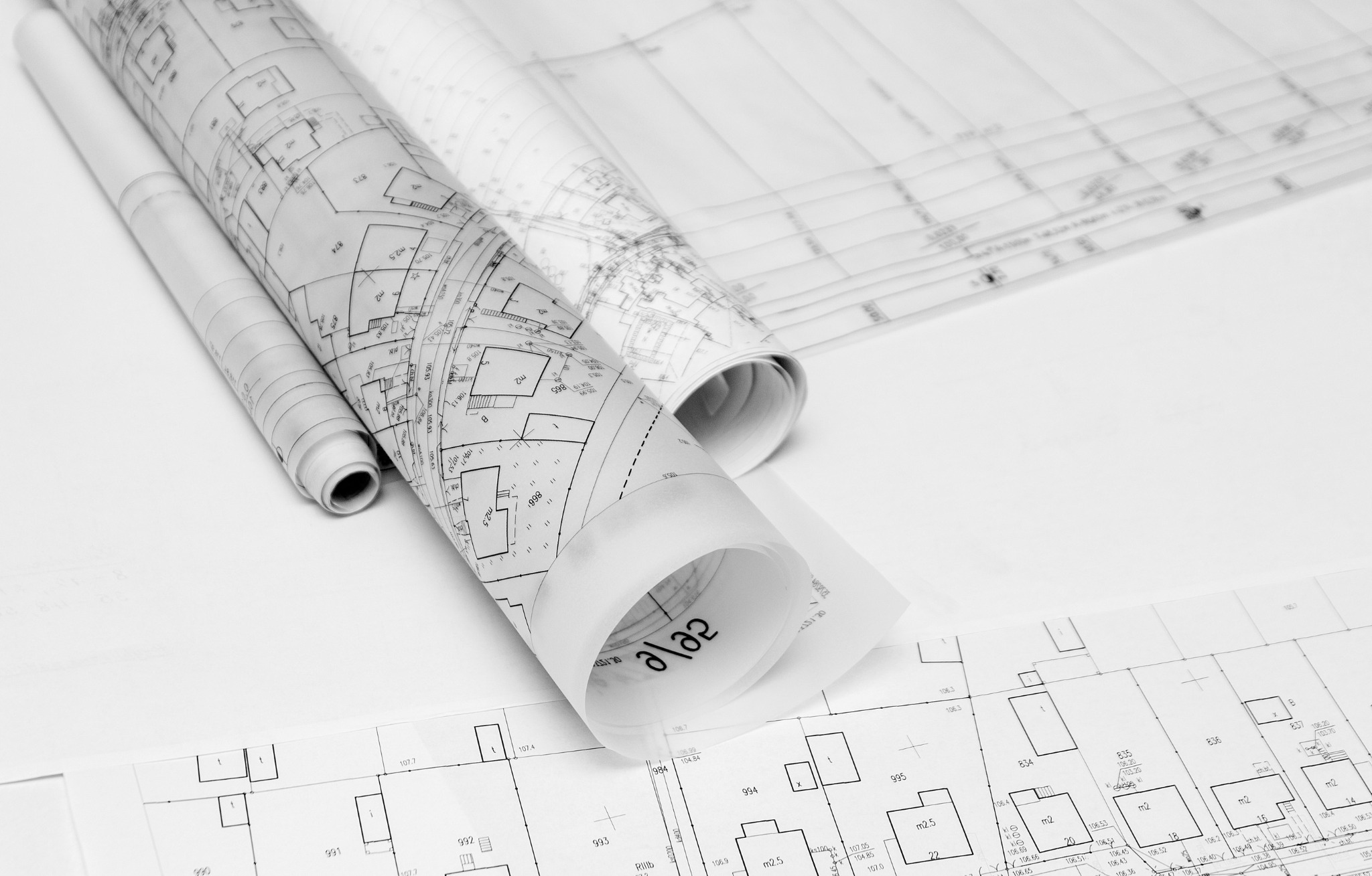 Architektoniczny plan budynku, częściowo zwinięty, z widocznymi detalami technicznymi i wymiarami, czarno-biały wydruk na białym tle.