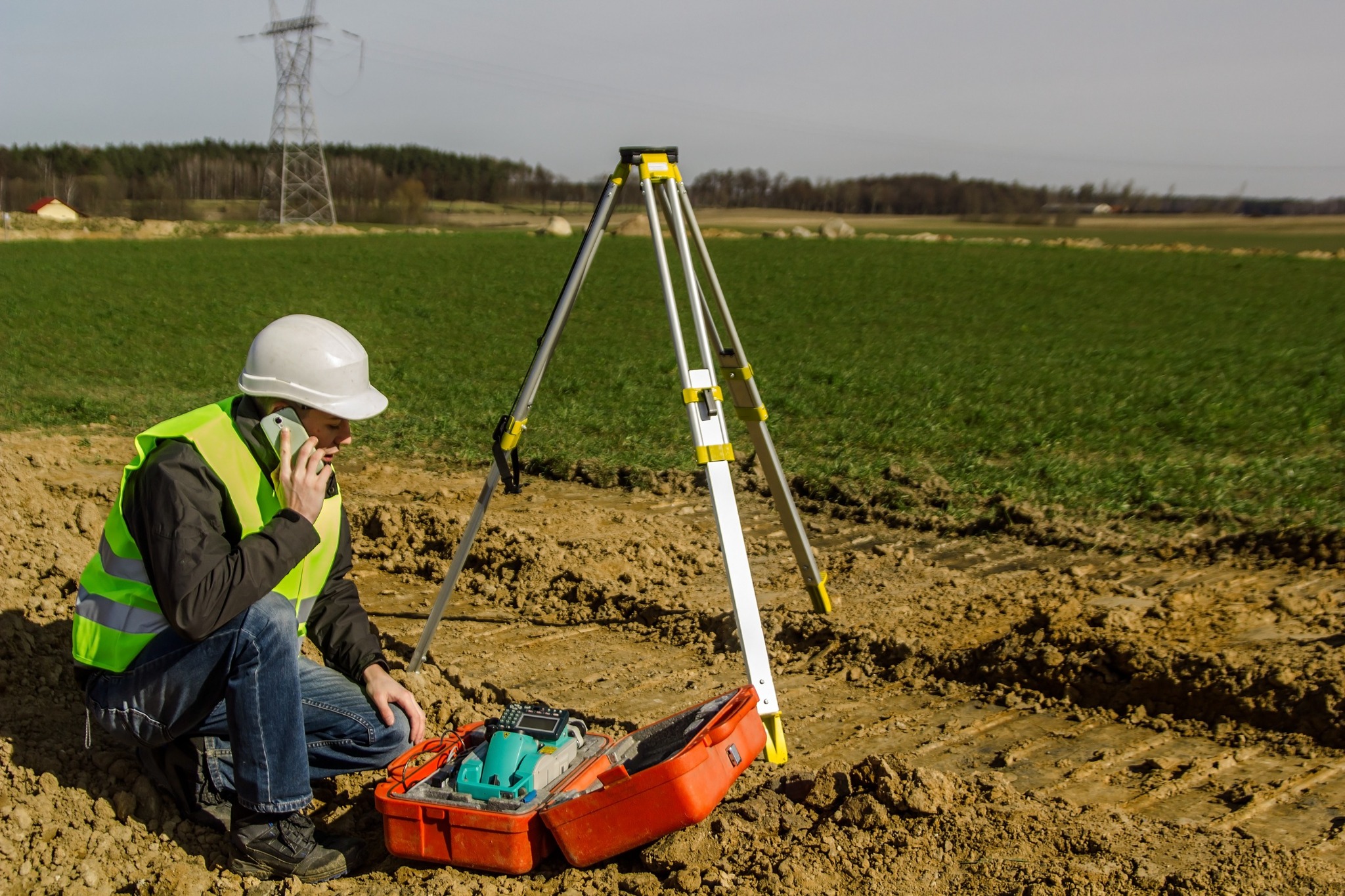 Geodeta w kasku i kamizelce odblaskowej rozmawia przez telefon obok niwelatora i otwartej walizki z urządzeniami pomiarowymi na tle pola i słupa wysokiego napięcia.