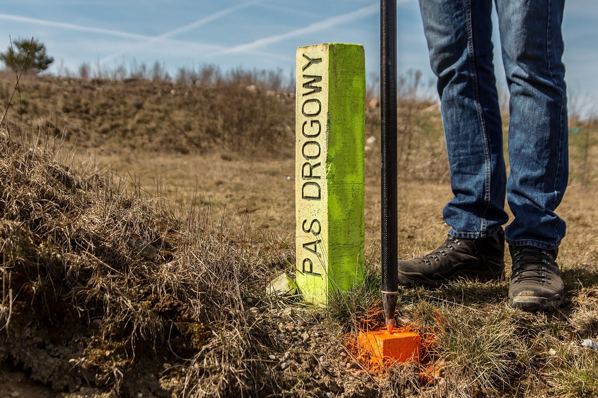 Żółto-zielony słupek graniczny z napisem 'PAS DROGOWY' wbity w ziemię obok pomarańczowego palika, w tle brązowa trawa i niebieskie niebo z białymi smugami kondensacyjnymi, obok stoi osoba...