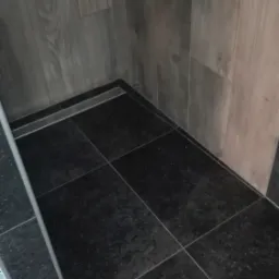 Ciemna, kwadratowa glazura podłogowa z metalowym odpływem liniowym w rogu łazienki w Gdyni, z widocznymi płytkami ściennymi imitującymi drewno.