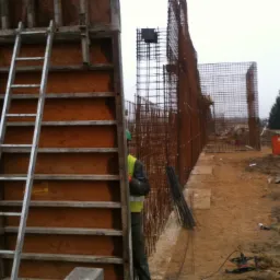 Przygotowanie zbrojenia i deskowania pod wylewanie betonu na placu budowy, widoczna drabina oparta o deskowanie, siatka zbrojeniowa i pracownik w kamizelce odblaskowej.