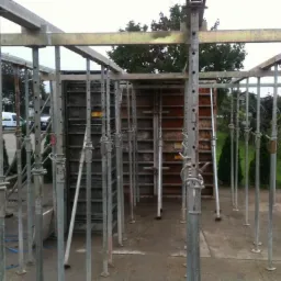 Konstrukcja szalunkowa z metalowymi podporami i deskami szalunkowymi w tle, przygotowana do wylewania betonu na fundamencie.