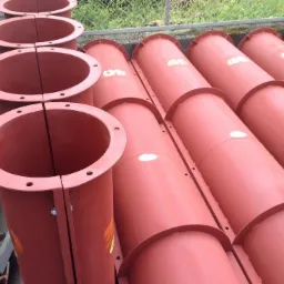 Rząd pomarańczowo-czerwonych, metalowych szalunków słupowych, okrągłych, z widocznymi otworami montażowymi i śrubami, ustawionych na zewnątrz, prawdopodobnie przed użyciem na budowie.