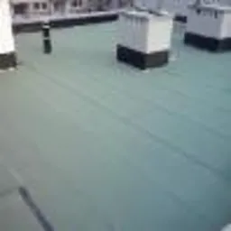 Zielona powierzchnia dachu pokryta papą termozgrzewalną, widoczne kominy wentylacyjne i fragmenty budynków w tle.