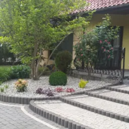 Ogród przed domem z krętą ścieżką z kostki brukowej, rabatą z roślin ozdobnych i drzew, schody prowadzące do wejścia.