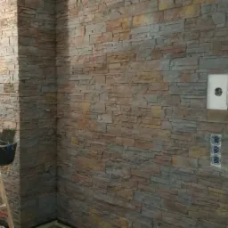 Ściana wyłożona dekoracyjną okładziną imitującą kamień, z widoczną drabiną i wiaderkiem, w trakcie prac wykończeniowych, z niezamontowanymi gniazdkami elektrycznymi.