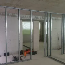 Montaż metalowej konstrukcji ścian działowych z płyt gipsowo-kartonowych w surowym wnętrzu, widoczne białe bloczki, narzędzia i drabina.