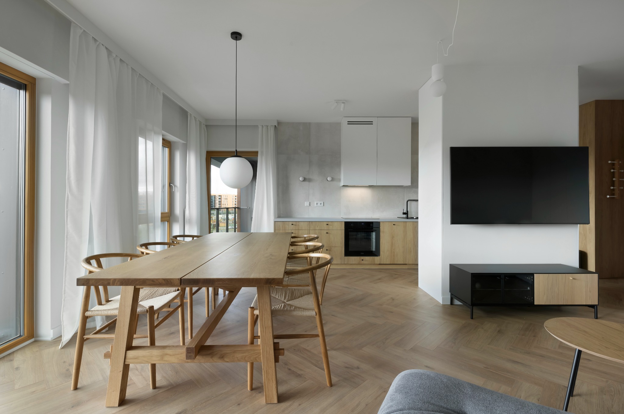 Jasne wnętrze z drewnianym stołem i krzesłami, parkiet w jodełkę, minimalistyczna kuchnia w tle. Nowoczesny design z dużym oknem i białymi zasłonami.