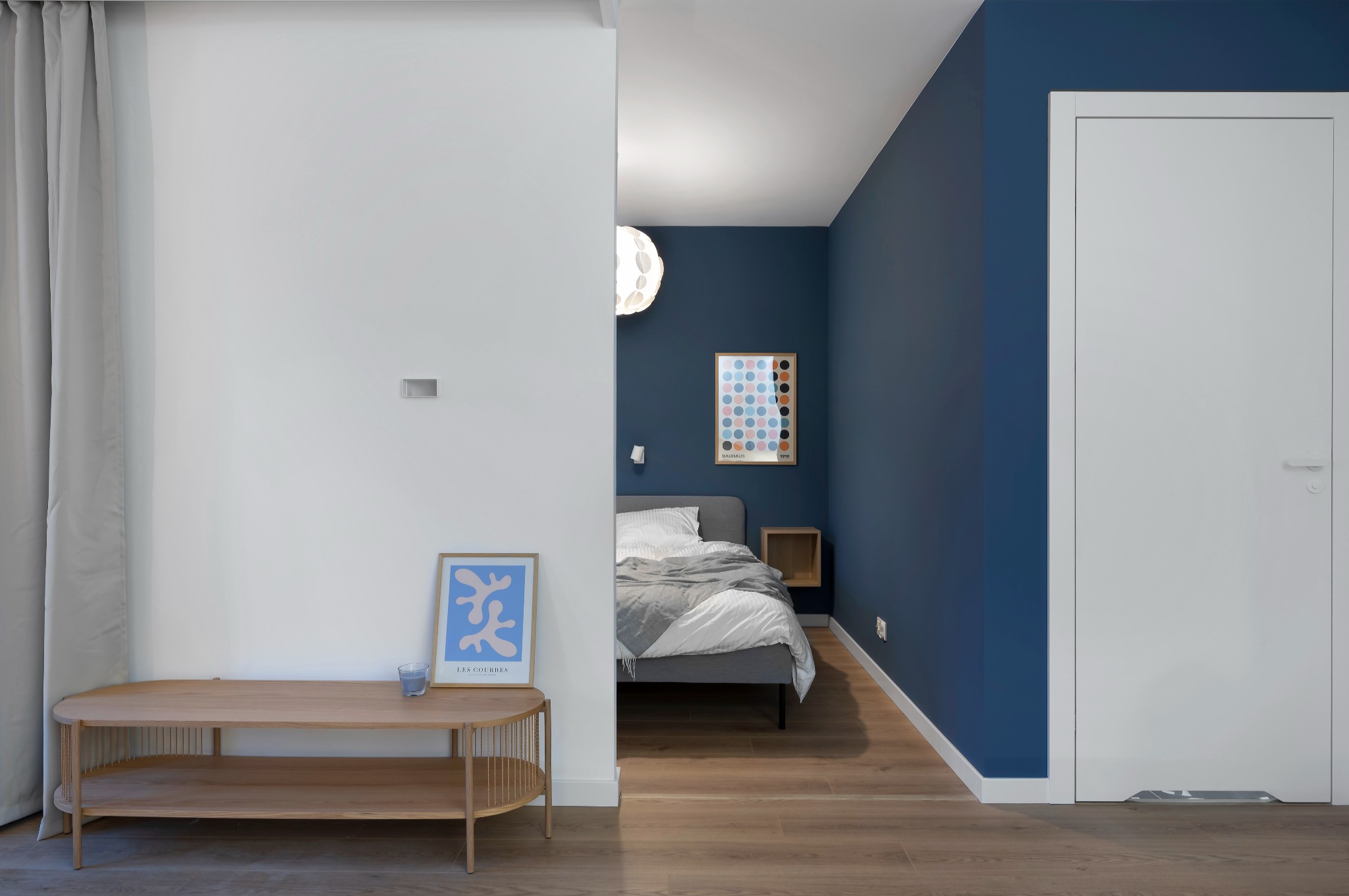 Nowoczesna sypialnia z akcentem granatowej ściany, drewnianą podłogą i minimalistycznym wystrojem. Widoczny fragment łóżka, stolik i plakat w ramie.