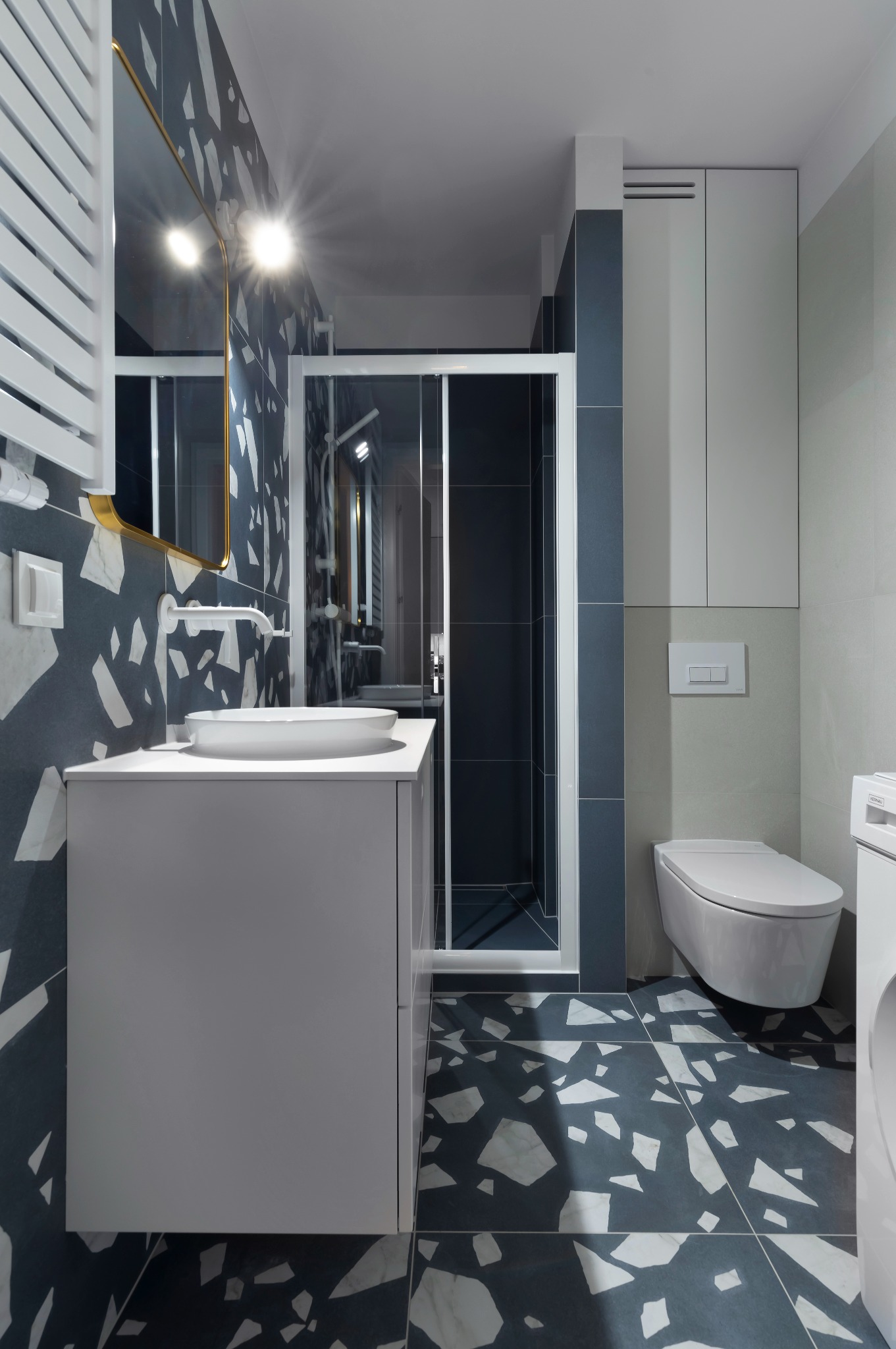 Nowoczesna łazienka z kabiną prysznicową, umywalką nablatową i płytkami w stylu lastryko. Złote akcenty lustra i minimalistyczny design.