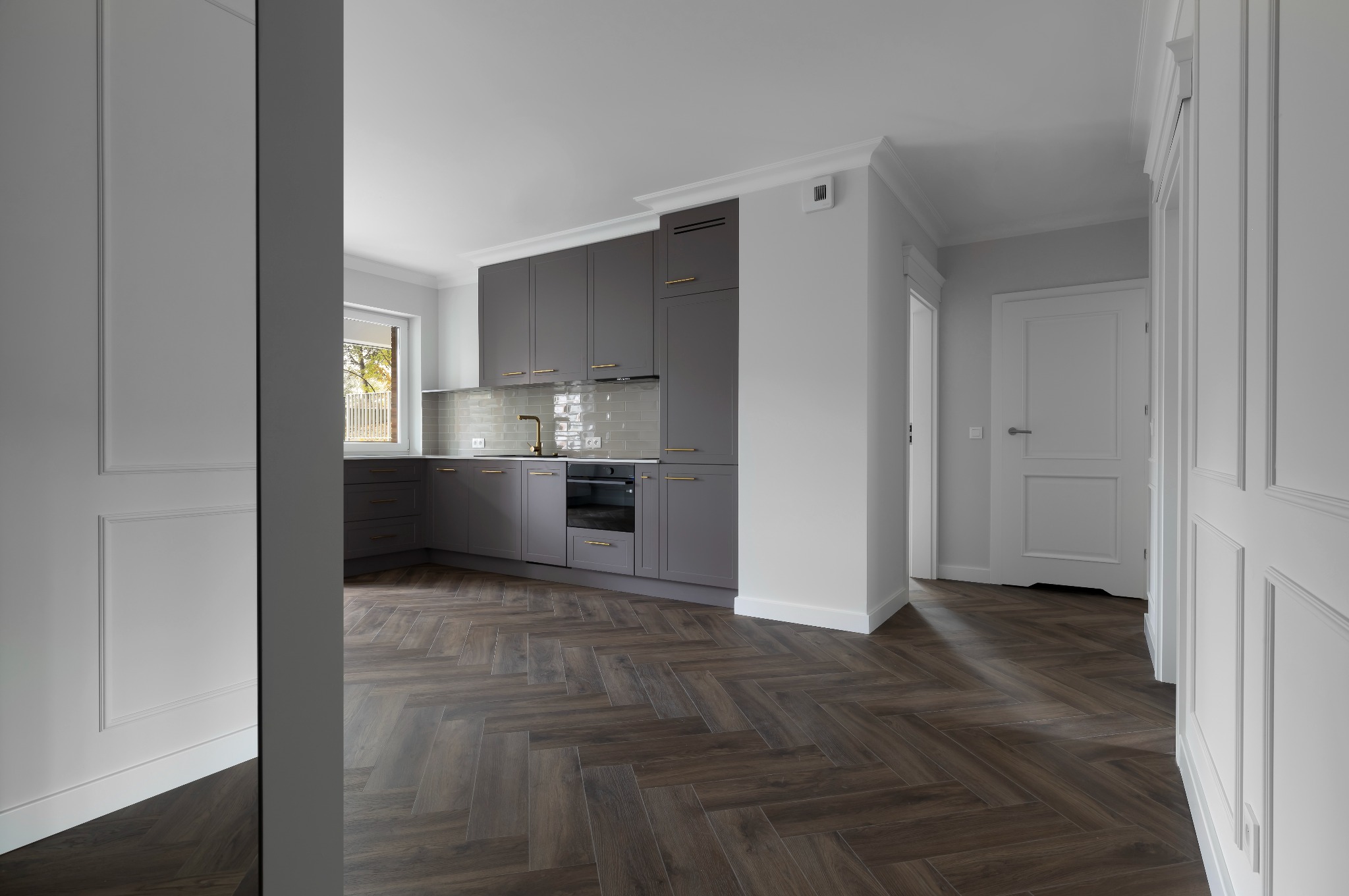 Szara kuchnia z parkietem w jodełkę, złote uchwyty, białe ściany z dekoracyjnymi listwami. Widok z korytarza, drzwi otwarte, okno w tle.