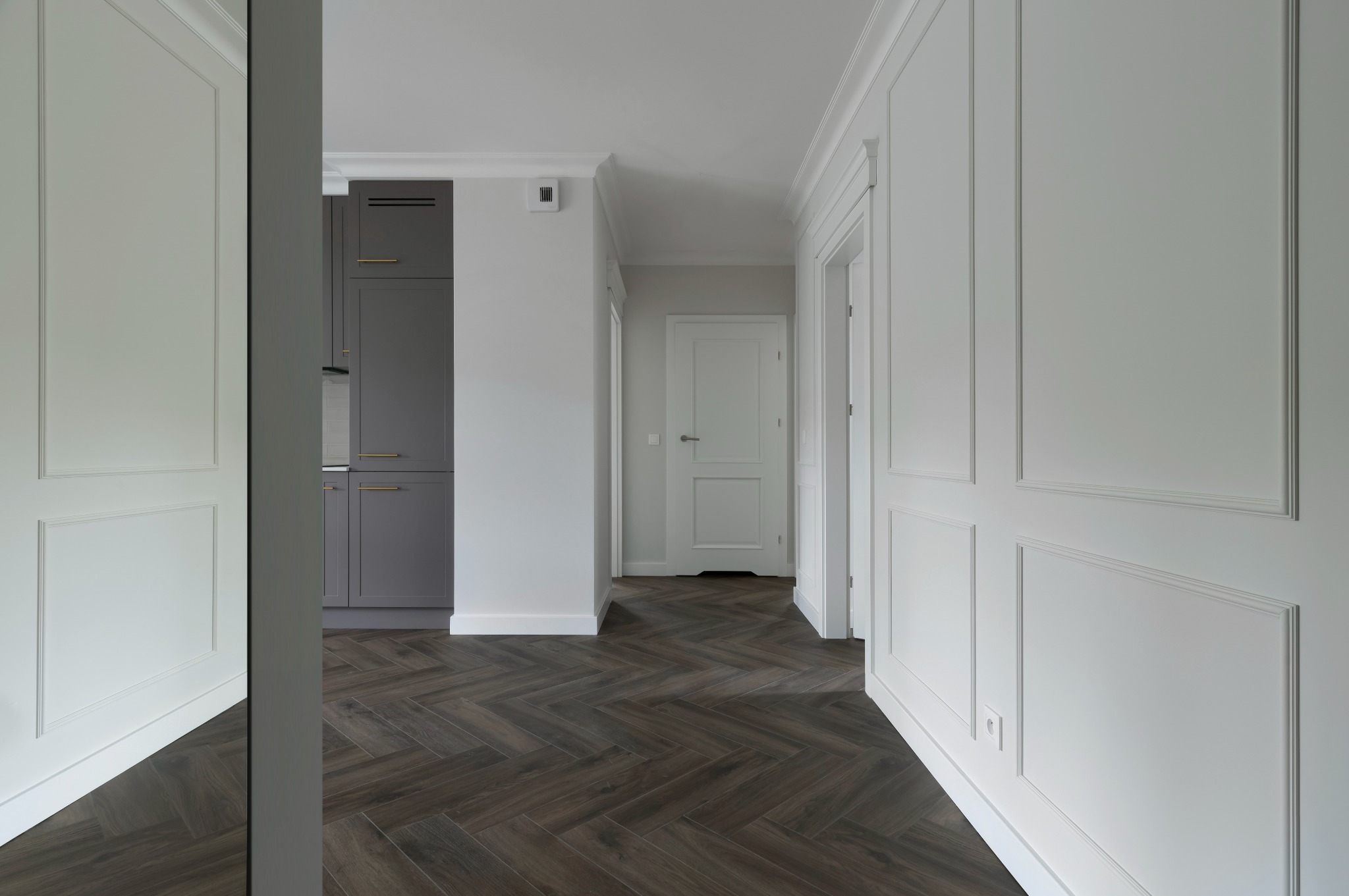 Eleganckie wykończenie wnętrza: białe ściany z dekoracyjnymi panelami, ciemna podłoga ułożona w jodełkę, widok na szarą zabudowę kuchenną przez lustro.