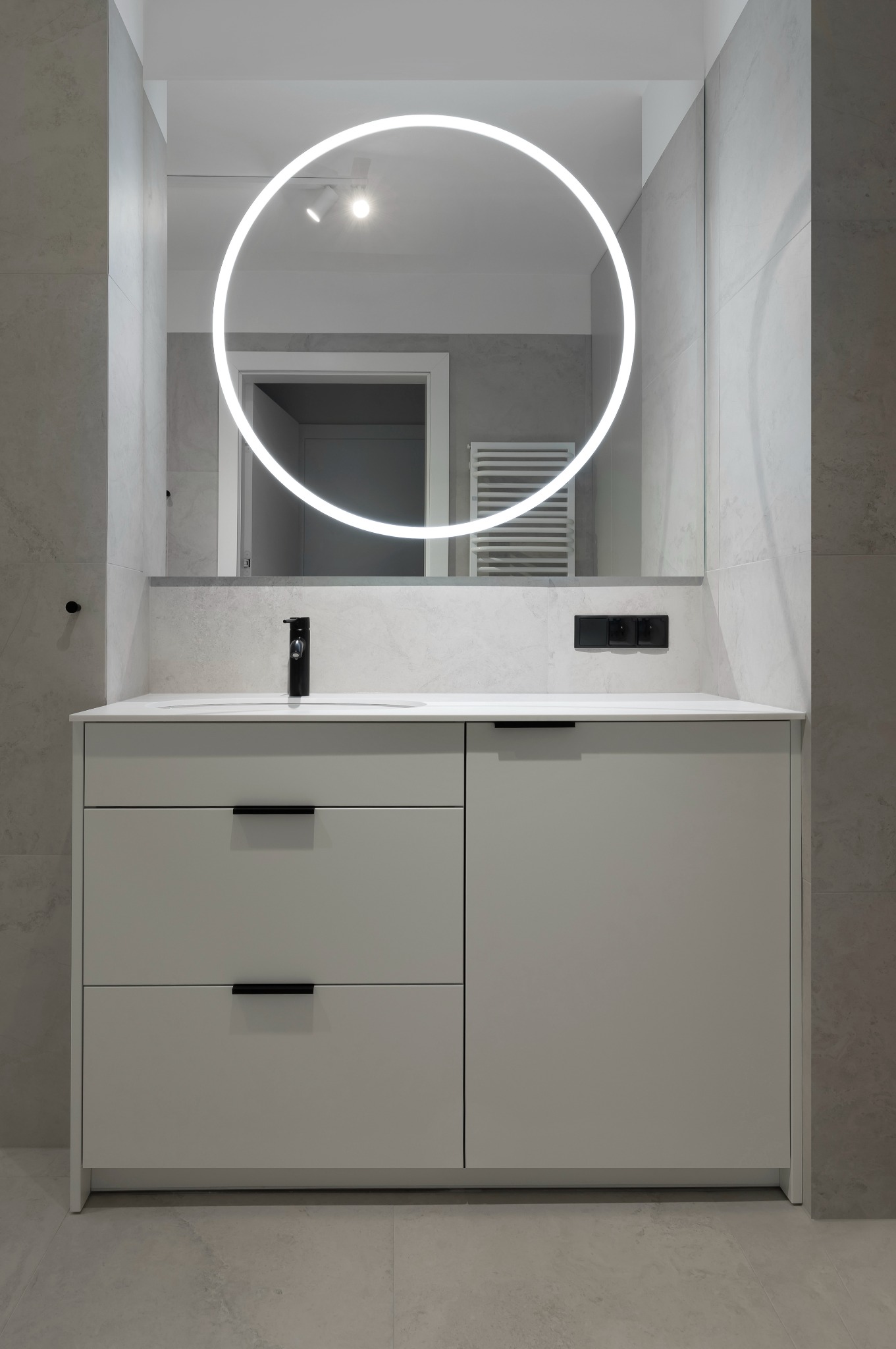 Nowoczesna łazienka z szarą szafką pod umywalkę, czarną armaturą i okrągłym lustrem z podświetleniem LED. Minimalistyczny design, płytki w odcieniach szarości.
