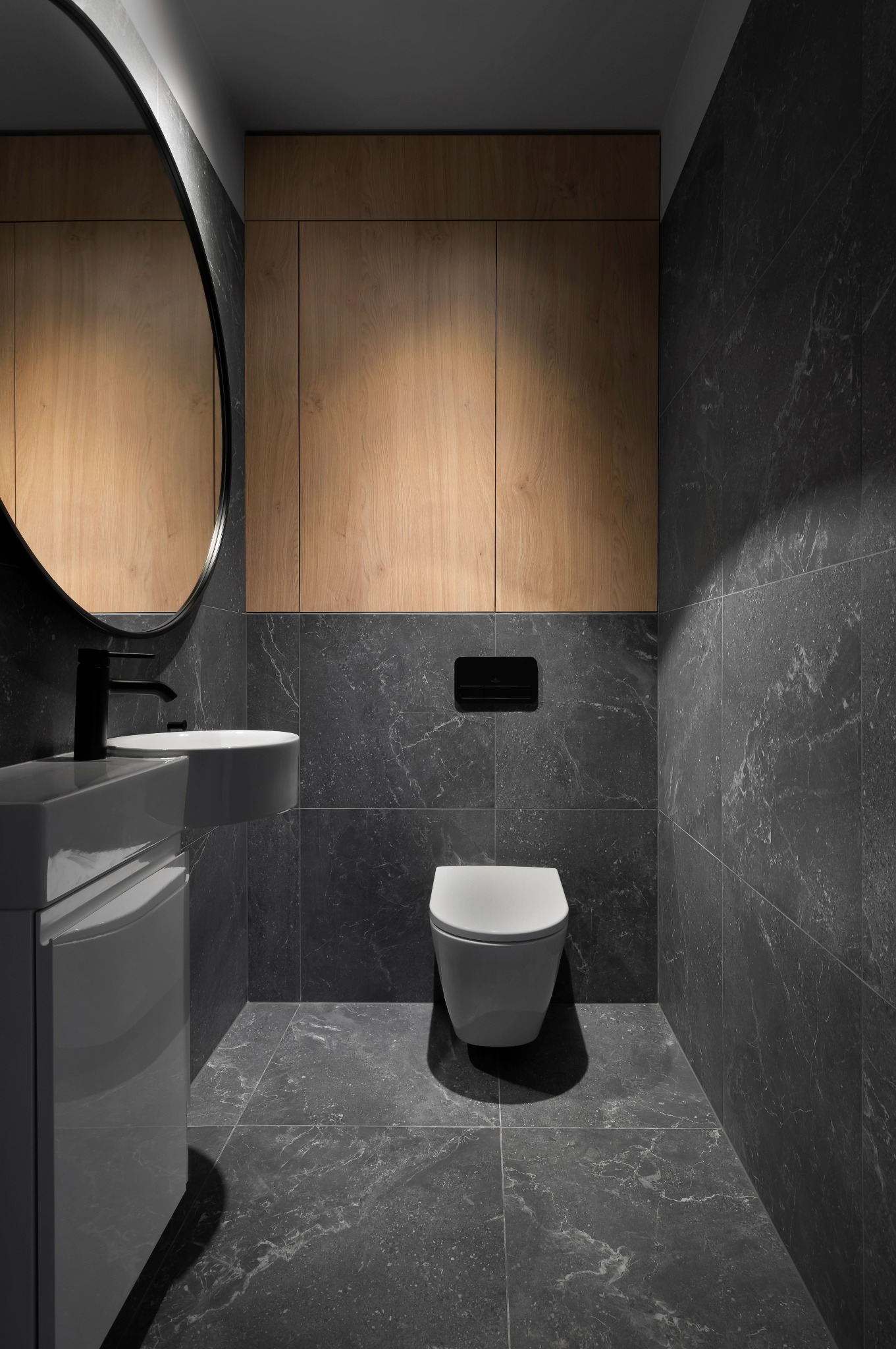 Nowoczesna łazienka z szarymi płytkami imitującymi marmur, wiszącą toaletą i umywalką nablatową. Drewniane szafki nadają ciepła minimalistycznemu wnętrzu.