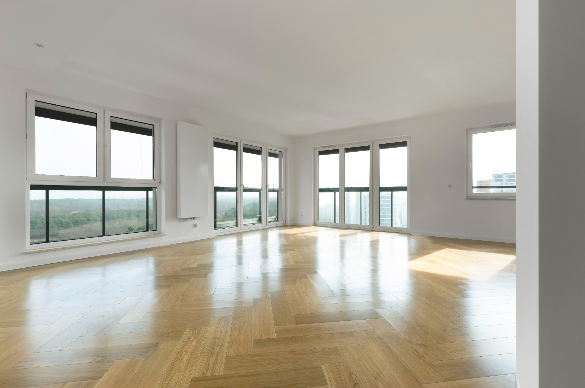 Jasne, przestronne wnętrze z parkietem w jodełkę, białymi ścianami i dużymi oknami z widokiem na zieleń. Minimalistyczny design, nowoczesne grzejniki.