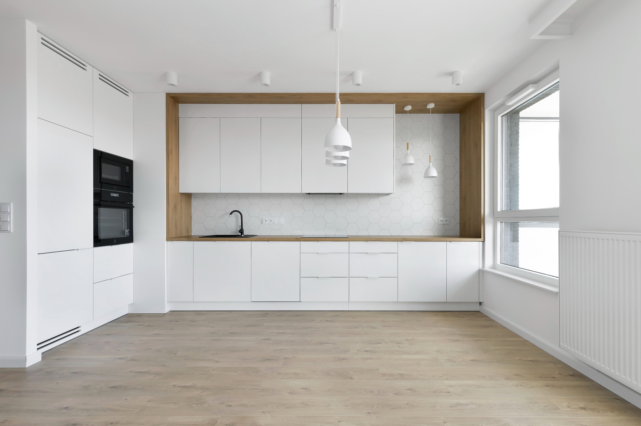 Nowoczesna, minimalistyczna kuchnia z białymi szafkami, drewnianym blatem i heksagonalnymi płytkami. Uwagę przykuwają designerskie lampy wiszące i wbudowane sprzęty AGD.