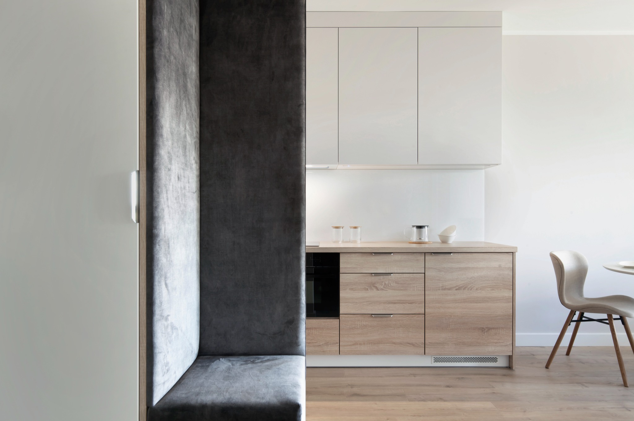 Nowoczesna kuchnia z drewnianymi szafkami i szarą, tapicerowaną wnęką do siedzenia. Minimalistyczny design, jasne kolory, drewniana podłoga.