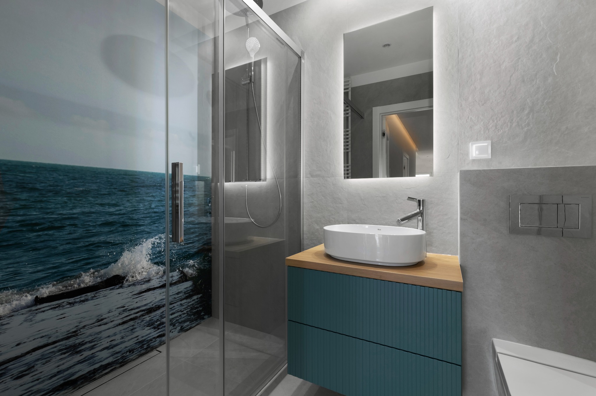 Nowoczesna łazienka z kabiną prysznicową z motywem morskim, umywalką nablatową na szafce z ryflowanym frontem i podświetlanym lustrem.