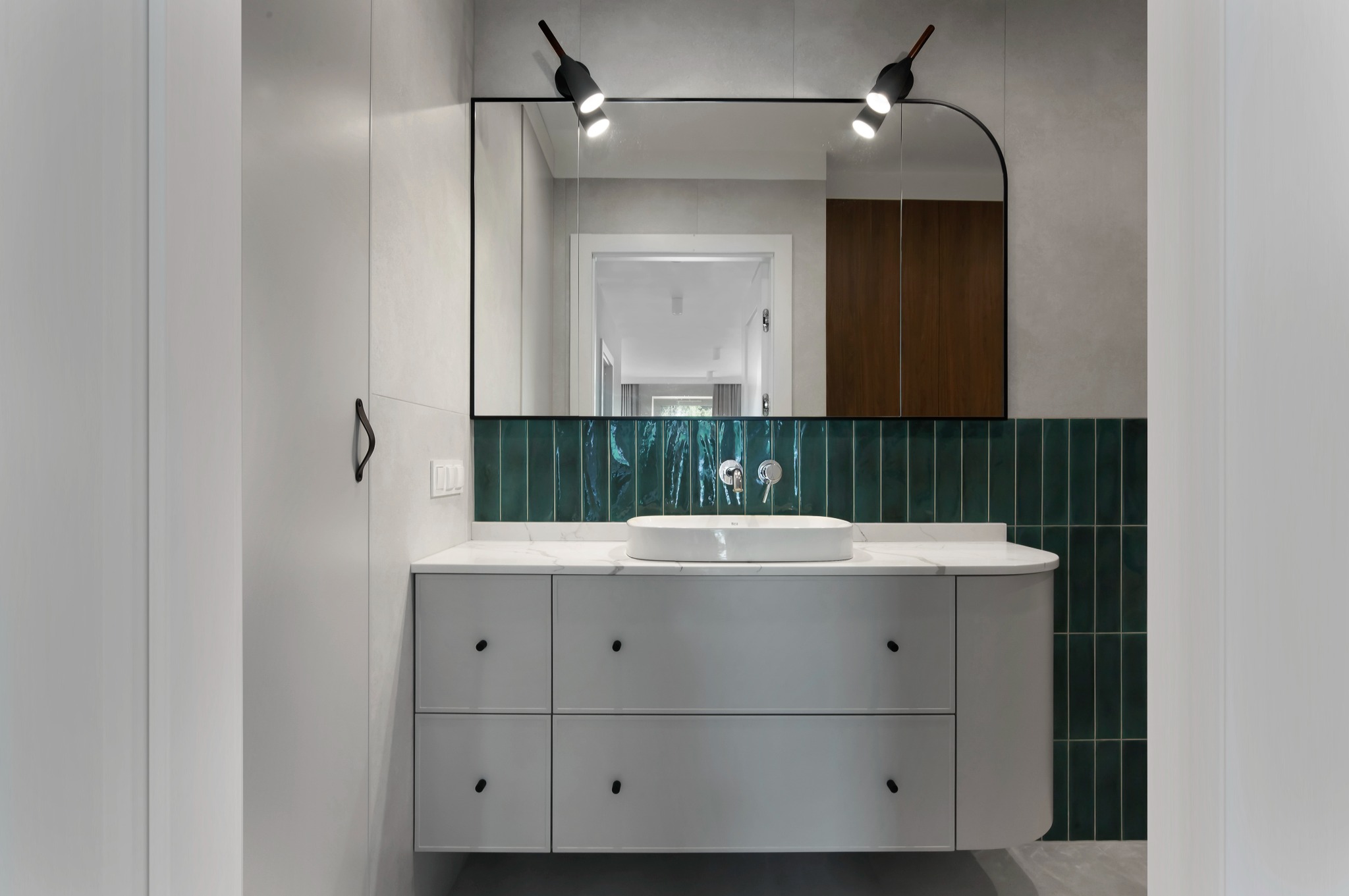 Nowoczesna łazienka z białą szafką pod umywalką, marmurowym blatem i zielonymi, błyszczącymi płytkami. Umywalka owalna, nad nią lustro z oświetleniem punktowym.