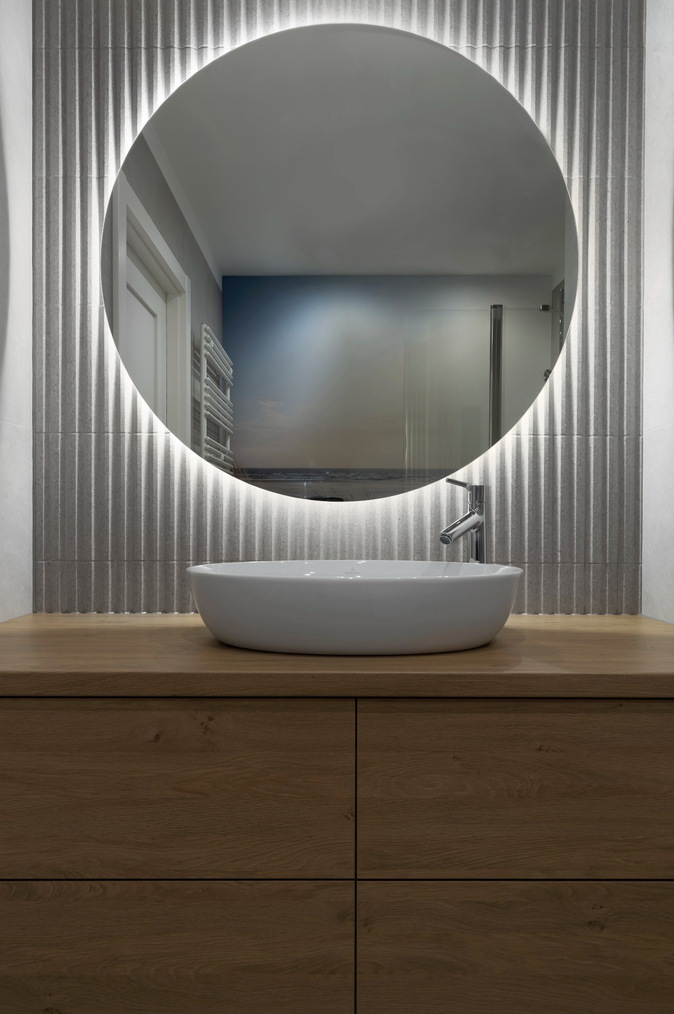 Nowoczesna łazienka: owalna umywalka nablatowa, drewniana szafka, podświetlane lustro na tle ściany z dekoracyjną fakturą. Minimalistyczny design.