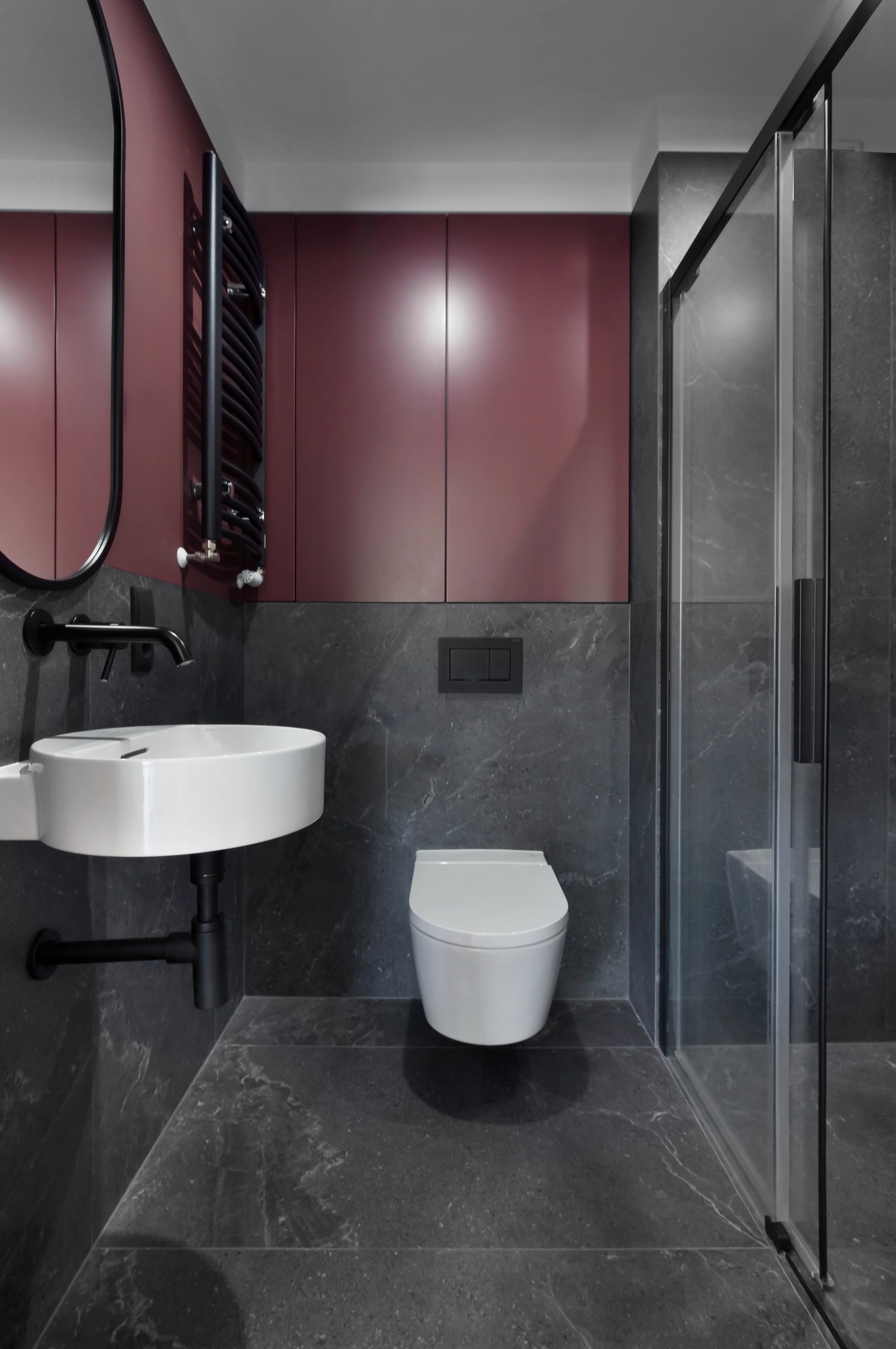 Nowoczesna łazienka z szarą marmurową okładziną, białą ceramiką i akcentami w kolorze burgundu. Kabina prysznicowa ze szklanymi drzwiami i czarnymi detalami.