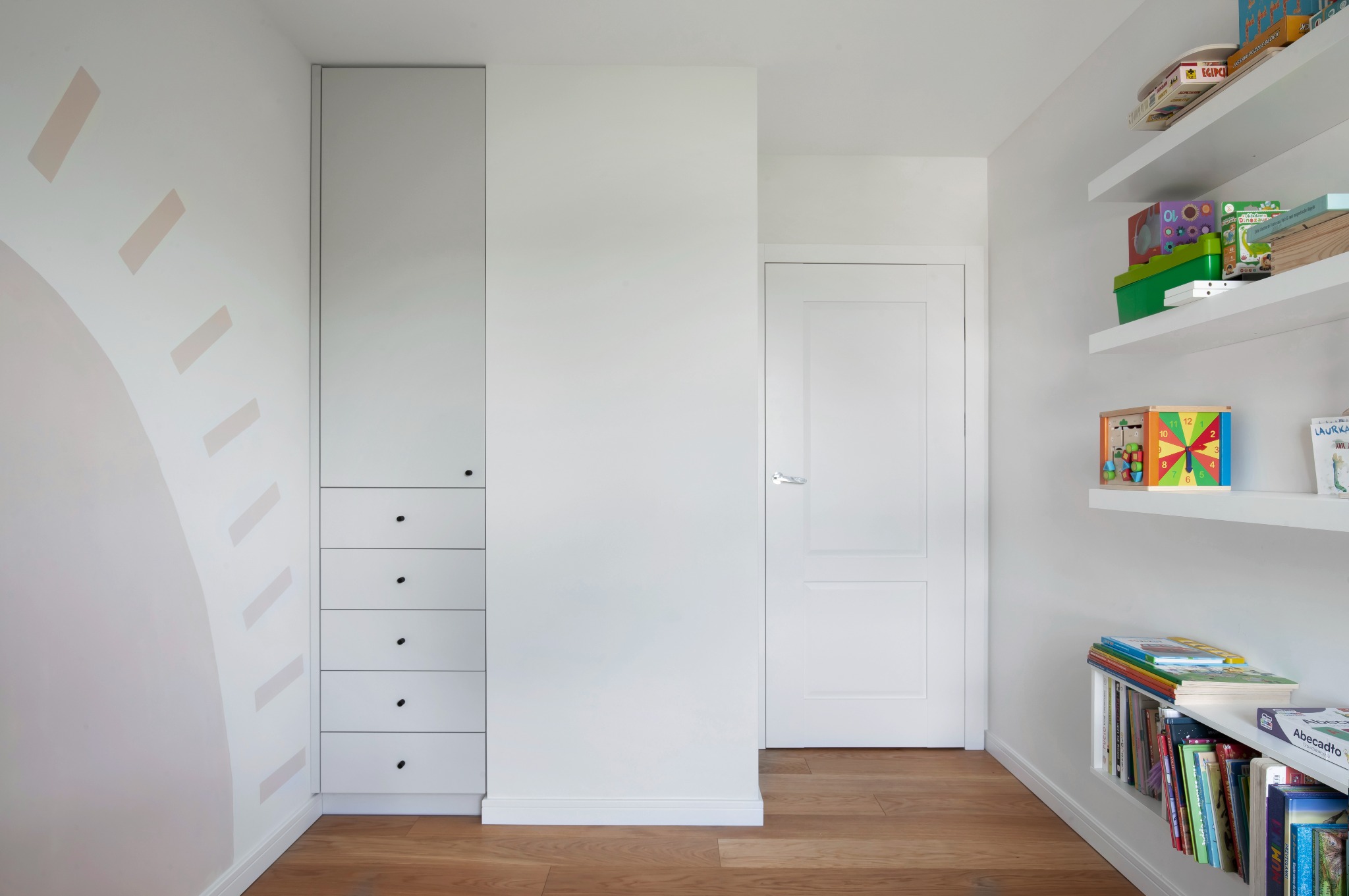 Pokój dziecięcy w bieli: zabudowa szafy z szufladami, drzwi i półki z zabawkami. Na ścianie dekoracja w kształcie słońca. Parkiet na podłodze.
