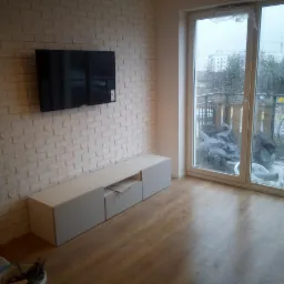 Salon po remoncie: ściana z białej cegły, telewizor na ścianie, biała szafka RTV, parkiet, okno balkonowe z widokiem na zewnątrz w deszczowy dzień.