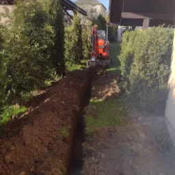 Wąski wykop w ziemi, wykonany minikoparką Kubota, pomiędzy tujami i budynkiem, na trawiastym terenie.