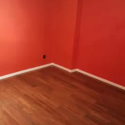 Pomarańczowy pokój z nową podłogą z paneli, białe listwy przypodłogowe i sufitowe, widoczny kontakt elektryczny na ścianie.