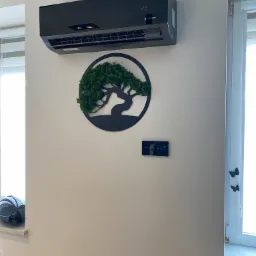 Czarny klimatyzator Vesser zamontowany na białej ścianie, pod nim dekoracja ścienna w kształcie drzewa z mchu, obok włączniki dotykowe i okno z dekoracyjnymi motylami.